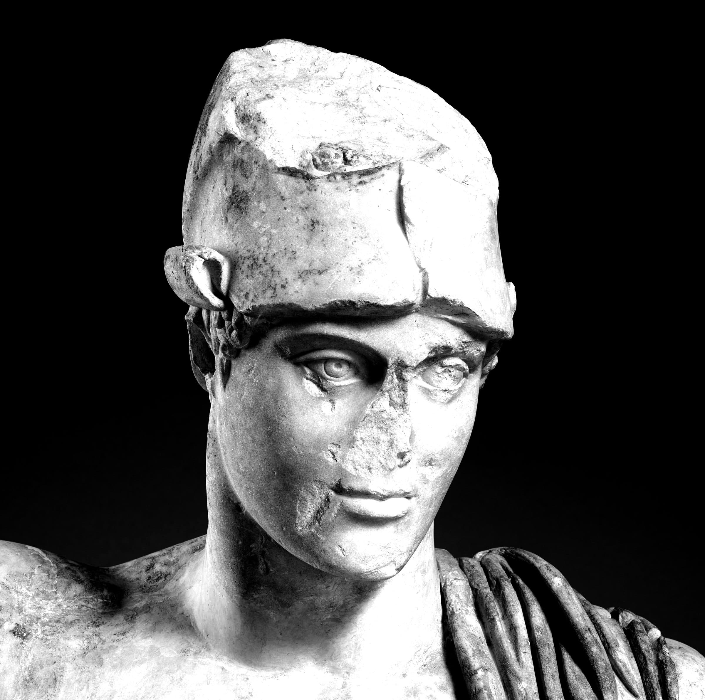 Marble head of a wounded Roman warrior: the helmet broken, the face weathered and cracked, the form intact